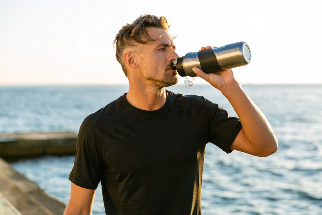 healthy adult man drinking water from bottle on seashore in front of sunrise